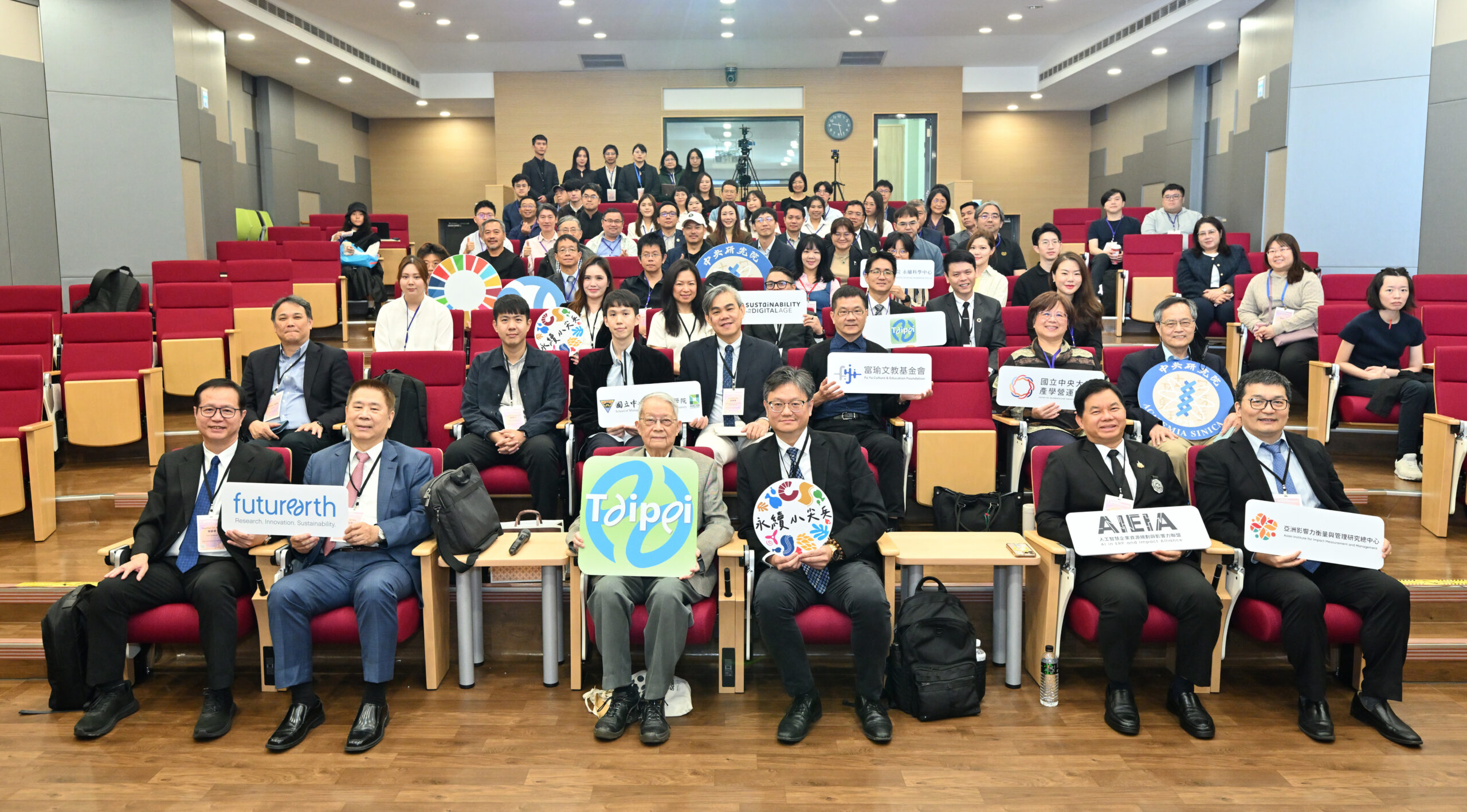 Conference participants at ICAISS2025 holding organizational signs and sustainability symbols in auditorium