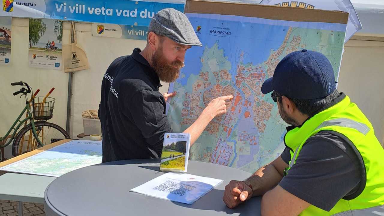 Two people discussing map of Mariestad at outdoor information booth with bicycle visible