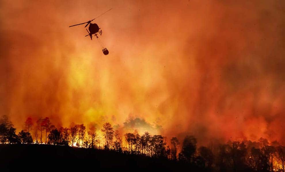 Helicopter with water bucket silhouetted against massive wildfire flames and smoke